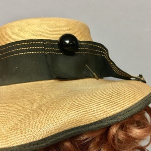May include: A straw hat with a wide brim, black ribbon, and a black button. The hat is trimmed with gold stitching.