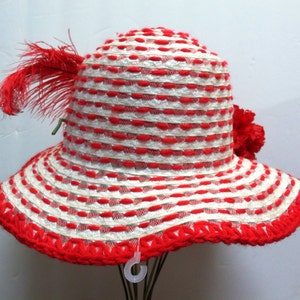Puede incluir: Un sombrero de sol a rayas rojas y blancas con un borde de ganchillo rojo y un adorno de plumas rojas.