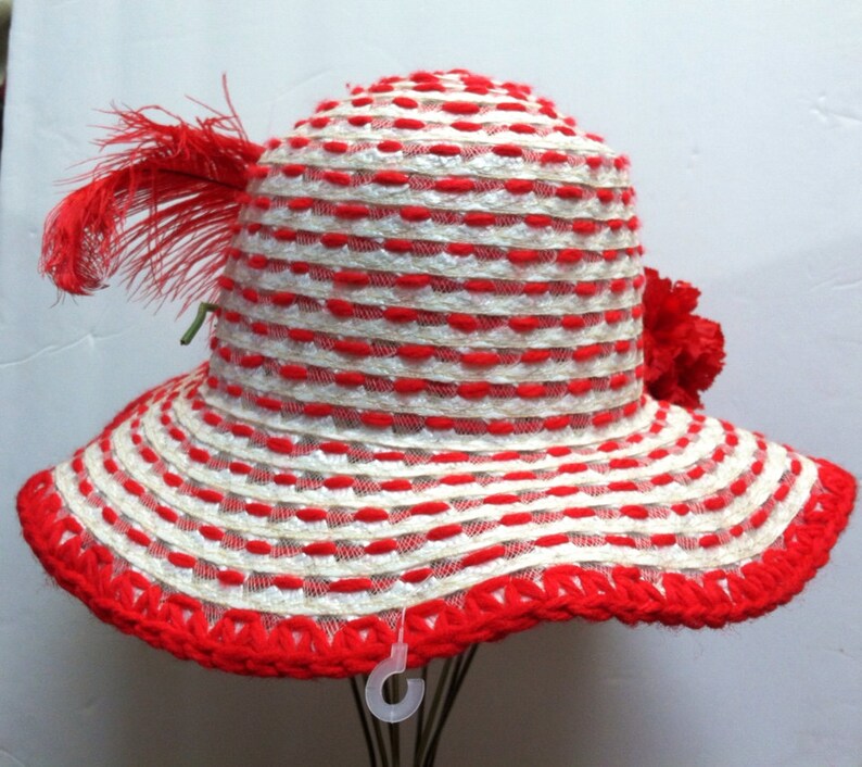 Puede incluir: Un sombrero de sol a rayas rojas y blancas con un borde de ganchillo rojo y un adorno de plumas rojas.