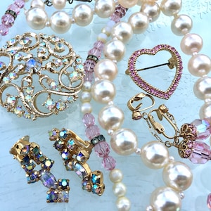 May include: A vintage jewelry set featuring a gold tone brooch with iridescent rhinestones, a heart-shaped brooch with pink rhinestones, and a pair of clip-on earrings with iridescent rhinestones. The jewelry is displayed on a strand of faux pearls and pink beads.