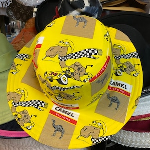 May include: Yellow bucket hat with a repeating pattern of camels wearing racing helmets and driving race cars. The hat has the text "Camel Filters" printed on it.