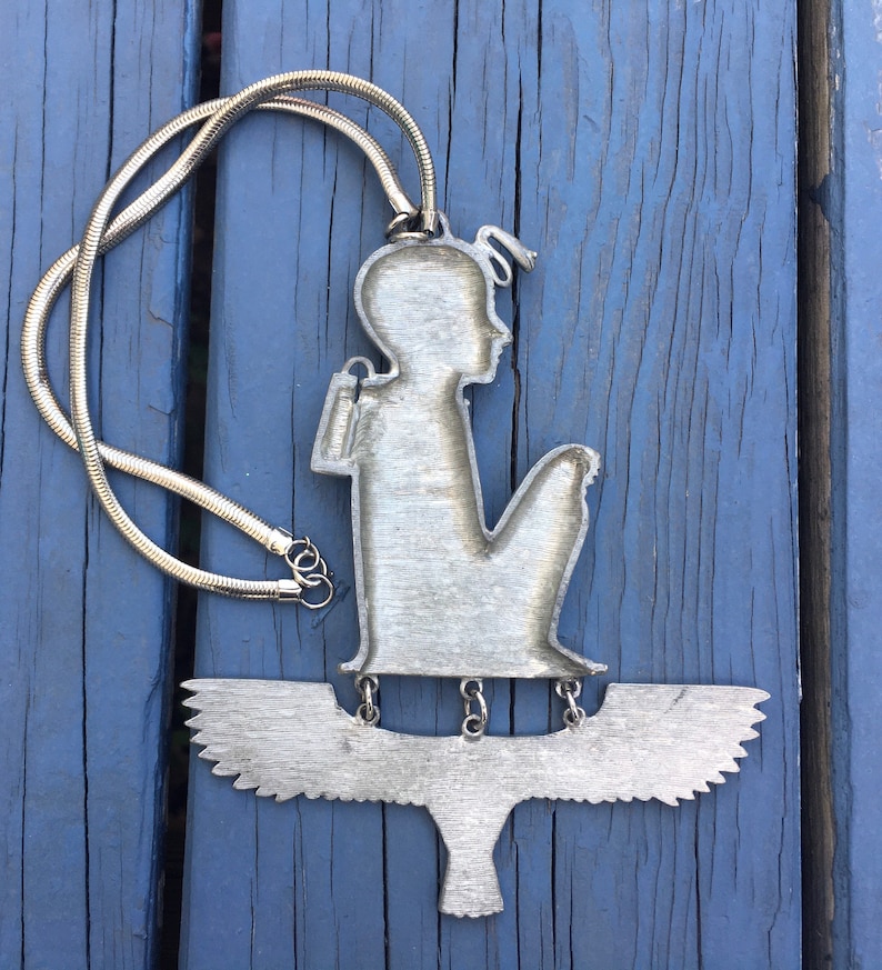 May include: Silver necklace with a pendant featuring a stylised figure of a person sitting on a bird with outstretched wings.