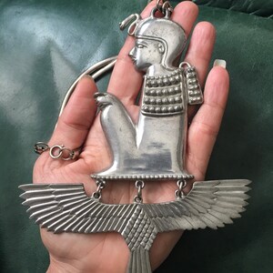 May include: Silver pendant necklace featuring a detailed depiction of a winged figure with a crown. The figure is seated with its arms crossed and has a detailed face and body. The wings are spread out and have a textured design.