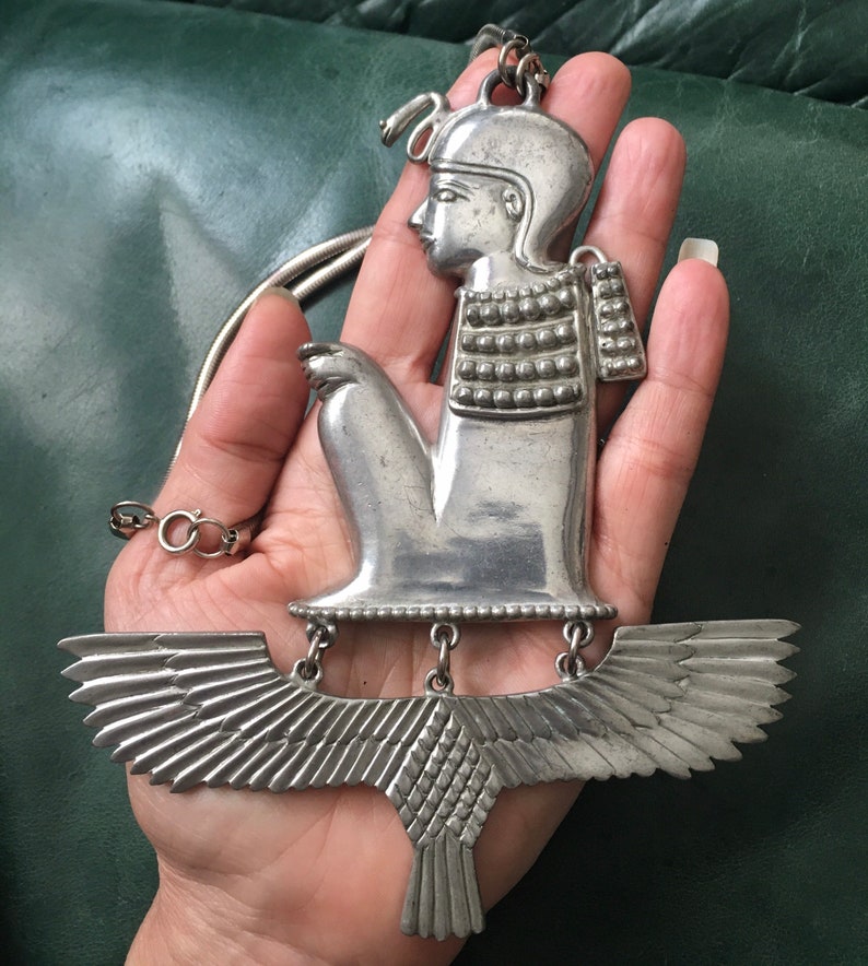 May include: Silver pendant necklace featuring a detailed depiction of a winged figure with a crown. The figure is seated with its arms crossed and has a detailed face and body. The wings are spread out and have a textured design.