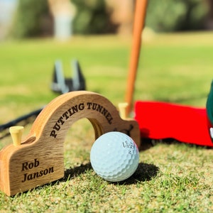 Peut inclure: Un tunnel de putting en bois avec les mots "Putting Tunnel" gravés sur le dessus. Le tunnel est personnalisé avec le nom "Rob Janson". Une balle de golf blanche repose au fond du tunnel.