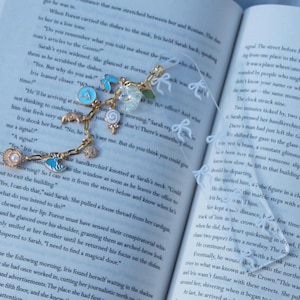 May include: A gold-toned bookmark with various charms, including a seashell, a blue butterfly, and a white flower, rests on an open book. The bookmark's design includes a delicate chain and a clear ribbon with small bow accents.