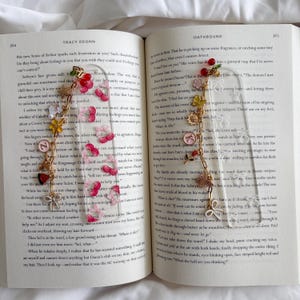 May include: Clear acrylic bookmark with gold chain and charms. The charms include red cherries, a gold sunflower, and a pink letter "K". The bookmark is placed on an open book.