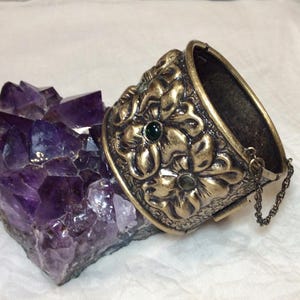 May include: A vintage gold-coloured hinged bracelet featuring floral patterns and a dark green gemstone. The bracelet rests on a large, purple amethyst crystal. A small chain is attached to the bracelet.
