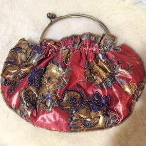 May include: A vintage red satin handbag with a floral pattern in gold, purple, and blue. The bag has a metal clasp and handle, and is decorated with beads. The design is reminiscent of a classic evening purse.
