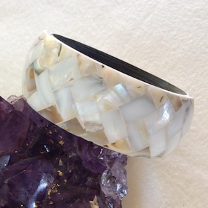 May include: A white and iridescent mother-of-pearl bracelet with a woven design. The bracelet is resting on a purple crystal. The interior of the bracelet is dark.