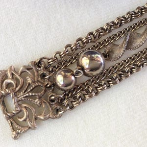 May include: Close-up of a vintage silver-toned bracelet. The bracelet features multiple strands of chain, decorative filigree elements, and round, polished beads. The design has an ornate, antique aesthetic.