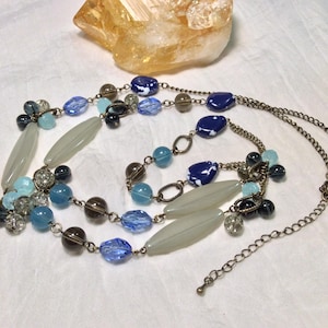 May include: A multi-strand necklace with blue, grey, and brown beads. The necklace has oval, round, and faceted beads, and a chain clasp. A yellow-orange crystal is in the background. The necklace is made of various beads.