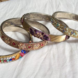 May include: A set of four metal bangle bracelets featuring various decorative elements. One bracelet has multicoloured rhinestones, another has floral patterns with red stones, one has purple accents, and the last has iridescent leaf-shaped beads.