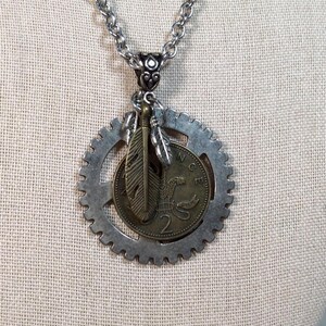 Peut inclure: Un pendentif en forme d'engrenage en métal argenté suspendu à une chaîne assortie. Le centre de l'engrenage contient une pièce de deux pence de couleur bronze et deux petites breloques en forme de plume. Le chiffre 2 et le texte PENCE sont visibles.