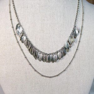 May include: A layered silver-tone necklace featuring two distinct chains. The shorter chain is adorned with a dense row of small, textured leaf-shaped charms, while the longer chain is a simple, delicate beaded chain with a smooth, polished finish.