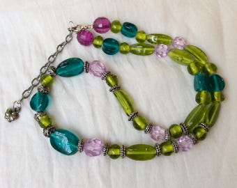 Puede incluir: Un collar de varias hebras con una variedad de cuentas translúcidas en tonos verde lima, verde azulado y morado claro. Las cuentas están separadas por pequeños espaciadores de metal plateado y el collar incluye una cadena extensora plateada.