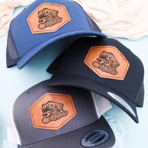 May include: Three trucker hats with a brown leather patch featuring a fish design and the text "On The Fly's". The hats are in blue, black, and gray.