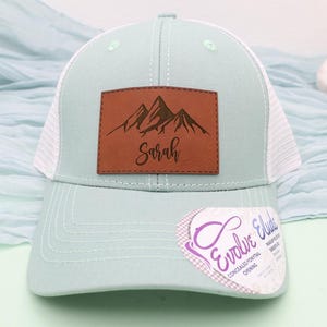 May include: A light blue trucker hat with a white mesh back and a curved brim. The hat features a brown leather patch with a mountain design and the name "Sarah" in script. A sticker is attached to the brim.