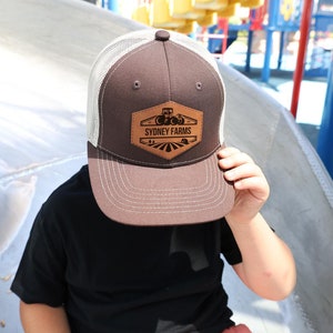 May include: A brown and white trucker hat with a brown leather patch that says "Sydney Farms" and a tractor graphic.