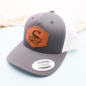 May include: A gray and white trucker hat with a brown leather patch featuring the letter "S" and the name "SMITH" in a stylized font.