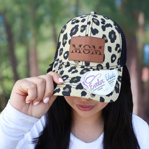 May include: A beige and black leopard print trucker hat with a black mesh back. The hat features a brown leather patch with the word "MOM" and the words "Rock, Emma, James" printed on it. The hat also has a white tag that says "Evolve Elude".