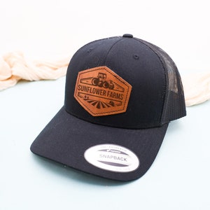 May include: Black trucker hat with a brown leather patch that says "Sunflower Farms" and a small image of a tractor. The hat has a black mesh back and a black snap closure.
