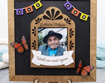 May include: Wooden memorial photo frame with a black background, featuring a photo of a man in a hat. The frame is decorated with orange butterflies, colorful bunting, and the text "Until we meet again."