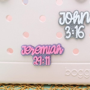 May include: A pink and white tote bag with the words "John 3:16" and "Jeremiah 29:11" written on it. The bag has a white handle and a white logo that says "BOGO" on the bottom.