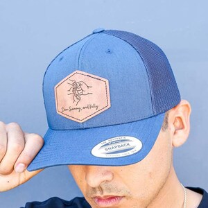 May include: A blue and black trucker hat with a brown leather patch featuring a bee and the text "Our Journey and Kelly".