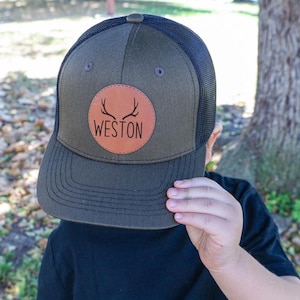 May include: A green and black trucker hat with a brown leather patch that says "WESTON" and has a pair of antlers inside the circle.