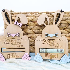 May include: Two wooden bunny money banks with slots for bills. The bunny on the left is pink and says "Natalie" and the bunny on the right is blue and says "Miguel".