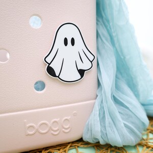 May include: A pink plastic tote bag with a white ghost sticker on the front. The tote bag has the word "bogg" printed on the bottom.