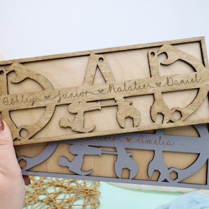 May include: Two wooden plaques with cut-out tool shapes and names. The top plaque is light brown and reads "DAD" with names Ashley, Junior, Natalie, and Daniel. The bottom plaque is gray and reads "Amelia".