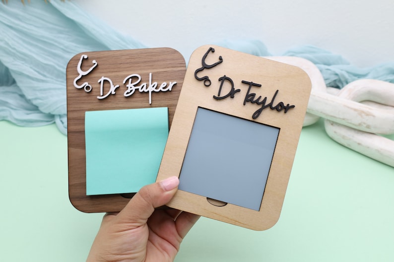 Pu&ograve; includere: Due porta note in legno con un design a stetoscopio e nomi personalizzati. Il porta note a sinistra ha una finitura in legno marrone chiaro e dice "Dr. Baker" con un post-it blu chiaro. Il porta note a destra ha una finitura in legno chiaro e dice "Dr. Taylor" con un post-it grigio.