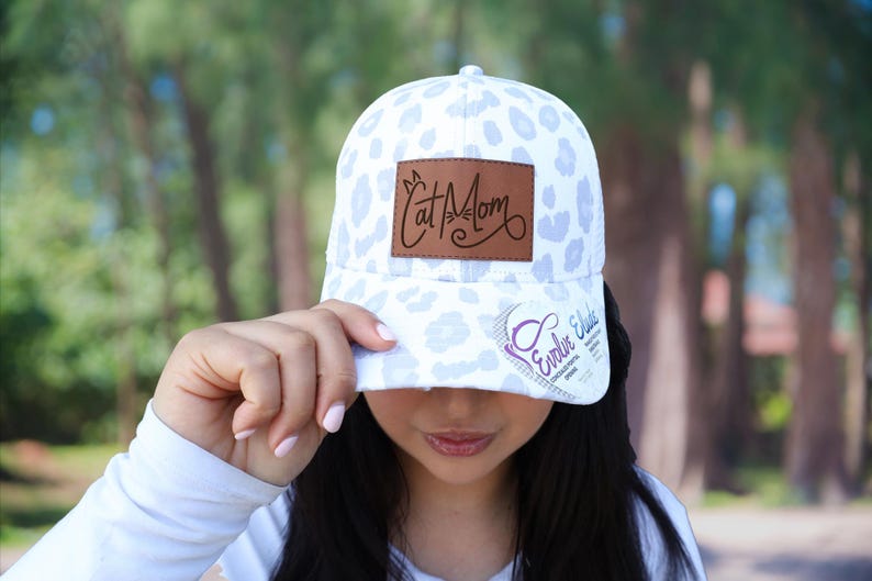 May include: A white baseball cap with a light blue leopard print pattern. The cap features a brown leather patch with the words "Cat Mom" and a logo on the side.
