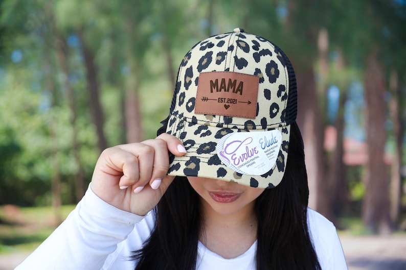 Pu&ograve; includere: Cappellino da camionista beige e nero con stampa leopardata e retro in rete nera. Il cappello presenta una toppa in pelle marrone con la scritta "MAMA" e "EST.2021" stampata sopra. Un adesivo bianco con la scritta "Evolve Elude" &egrave; attaccato alla visiera.