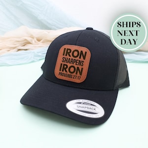 May include: Black trucker hat with a brown leather patch that reads "IRON SHARPENS IRON PROVERBS 27:17" in white lettering. The hat has a curved brim and a mesh back. The hat has a snapback closure.