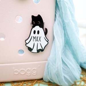May include: A black cat wearing a white ghost costume with the name "Max" written on it. The costume is attached to a pink and white tote bag.