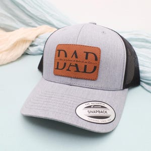 May include: A gray trucker hat with a black mesh back and a brown leather patch. The patch features the word "DAD" in large, bold letters, with additional text below. The hat has a curved brim and a snapback closure.