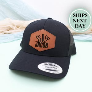 May include: Black baseball cap with a brown leather patch featuring barber tools and the name "JACOB". The cap has a curved brim, a mesh back, and a snapback closure. The hat is a Yupoong Snapback.
