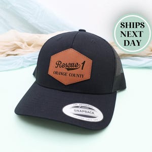 May include: Black trucker hat with a brown leather patch that reads "Rescue 1 Orange County". The hat has a curved brim and a mesh back. The hat has a Snapback tag.