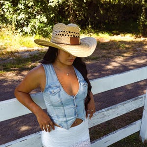 May include: A woman wearing a light blue denim vest with a white top and a straw cowboy hat with a brown leather band. She is standing in front of a white wooden fence.