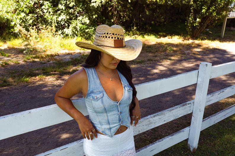 May include: A woman wearing a light blue denim vest with a white top and a straw cowboy hat with a brown leather band. She is standing in front of a white wooden fence.