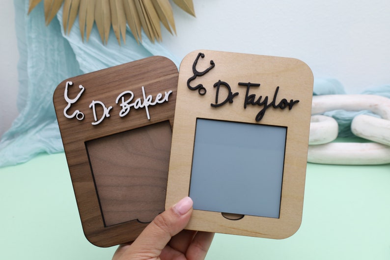 Pu&ograve; includere: Due cornici per foto in legno con un simbolo di stetoscopio e il testo "Dr. Baker" e "Dr. Taylor" su di esse. Le cornici sono entrambe di colore marrone chiaro e hanno una piccola apertura per una foto.