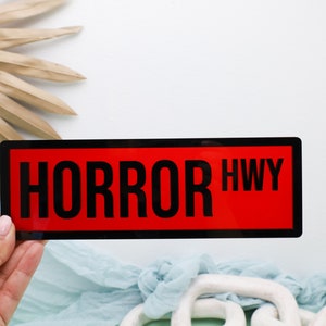 May include: A black and red rectangular sign with the text "HORROR HWY" in white.