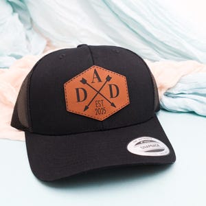 May include: Black baseball cap with a brown leather patch that reads "DAD" with crossed arrows and "EST 2025". The cap has a mesh back and a curved brim. A Snapback sticker is on the brim.