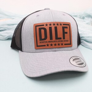 May include: A gray trucker hat with a black mesh back and a brown rectangular patch. The patch features the word "DILF" in bold black letters, with the words "DEVOTED. INVOLVED. LOVING. FATHER" below. The hat has a curved brim.
