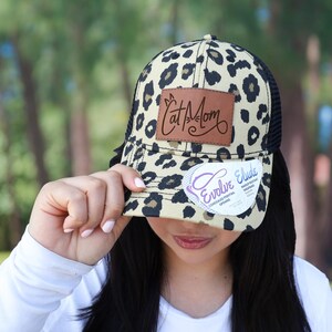 May include: A beige and black leopard print trucker hat with a black mesh back. The hat features a brown leather patch with the words "Cat Mom" and a white tag that says "Evolve Elude".