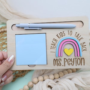 May include: Wooden desk organizer with a light blue pen, a blue sticky note pad, and a rainbow design with the text "I teach kids to talk back" and "Ms. Peyton". The organizer is made of light-colored wood.