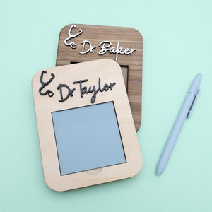 Pu&ograve; includere: Due cornici in legno con un'icona di stetoscopio e il testo "& Dr. Baker" e "& Dr. Taylor" su uno sfondo verde chiaro. C'&egrave; anche una penna blu chiaro nell'immagine.